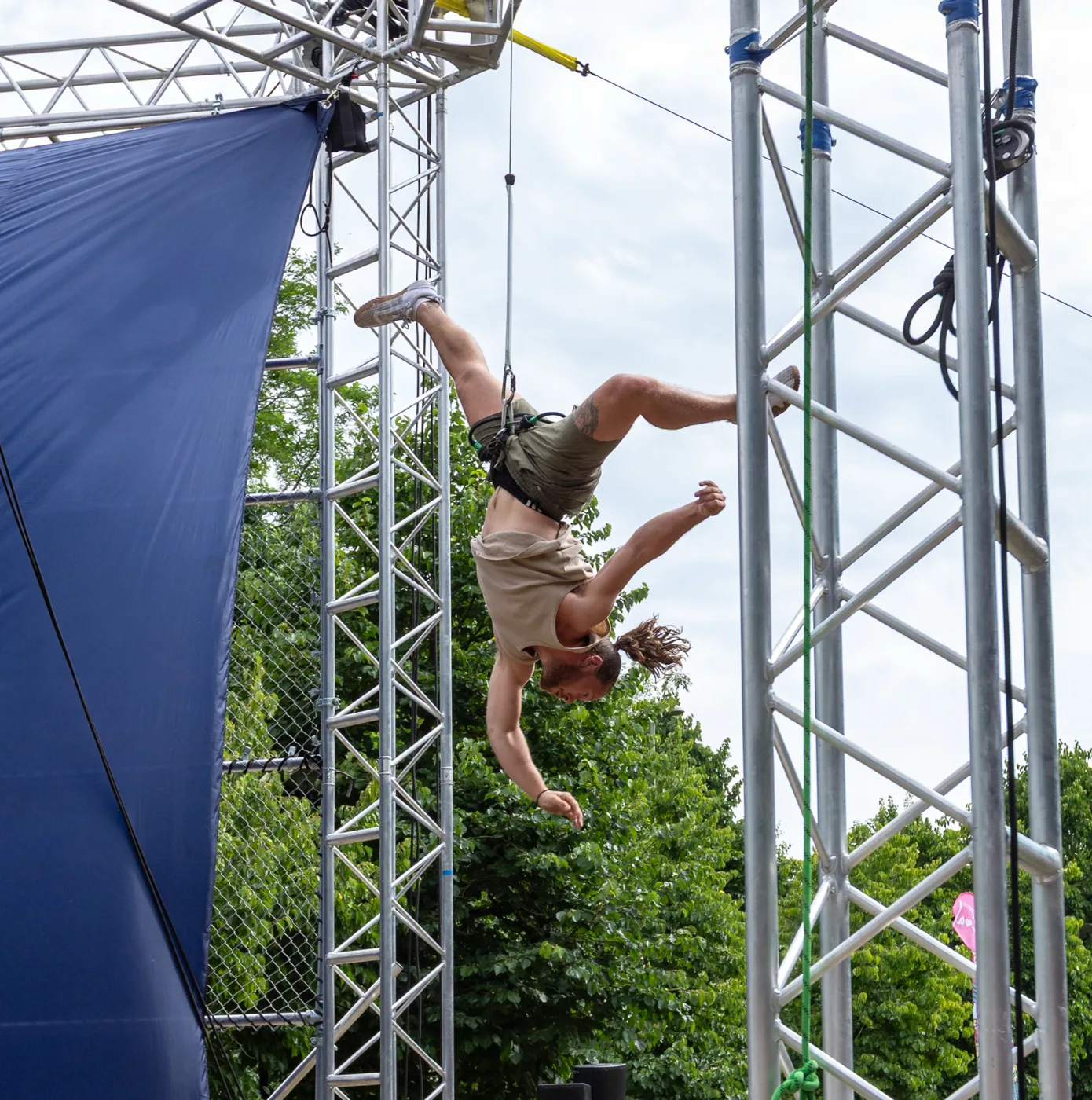 A performer hanging upside down from a harness beneath the circular metal rig, arms outstretched, with blue and purple fabric ribbons